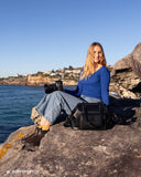 Woman sitting on a rock by the ocean with a camera and Noir Mirage Camera Bag. Stylish and functional camera bag